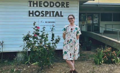 A woman in a dress and boots stands in front of a building which is labelled Theodore Hospital